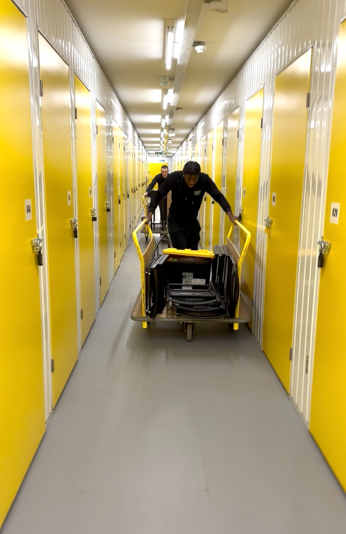 Two team members working together in bright yellow storage facility corridor with professional trolley